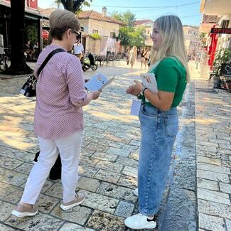 The image shows a woman handing a pamphlet to a woman dressed in a pink shirt. They are standing on a cobblestone street in front of a building. There are other people in the background. The woman in pink looks at the pamphlet and holds it in her hands. The woman holding the pamphlet is smiling. They appear to be engaged in conversation.