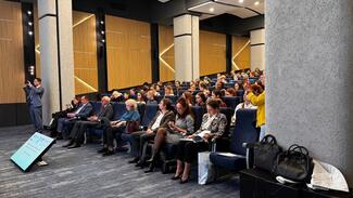Attendees sitting in a hall, watching and listening to a speaker during an event.
